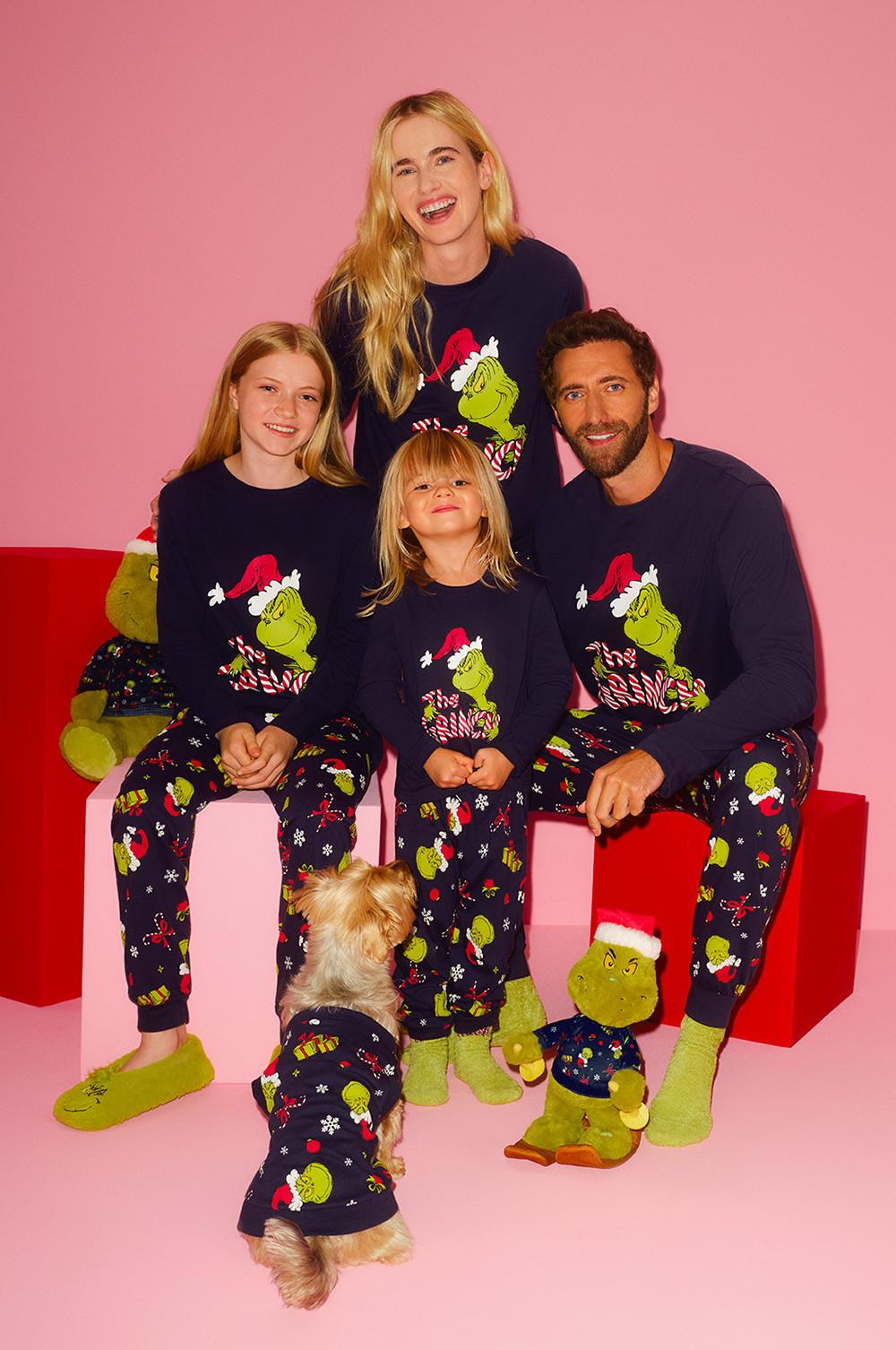 Family wearing matching Grinch Christmas pyjamas with a small dog in coordinating outfit against pink background.