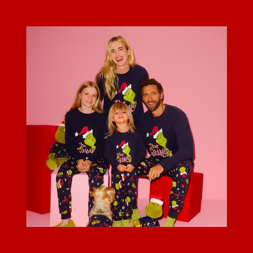 Family wearing matching Grinch Christmas pyjamas with navy tops and patterned bottoms, posed together with a small dog.
