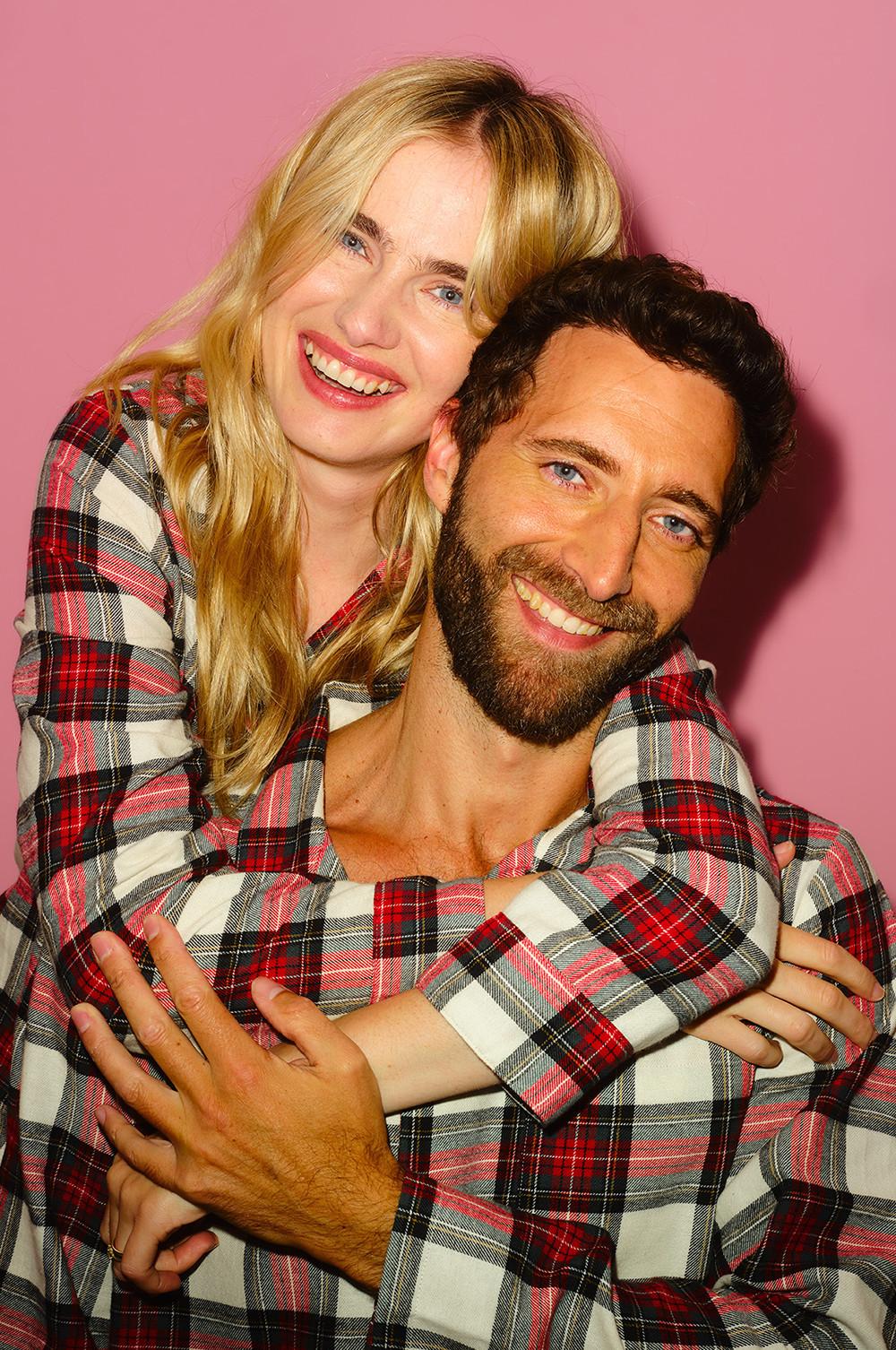 Two smiling people wearing matching red and white plaid flannel shirts embracing against a pink background.