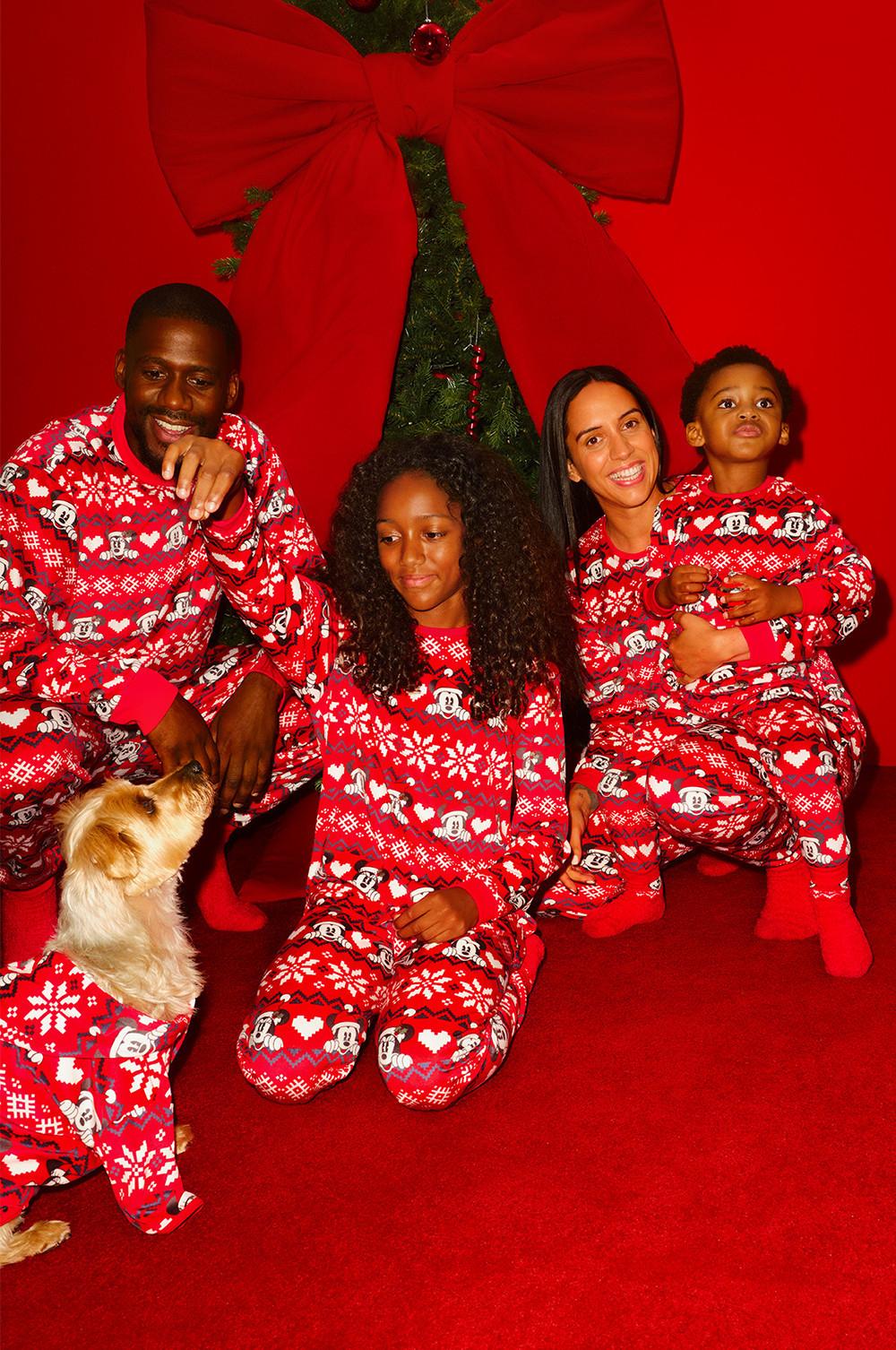 Family wearing matching Mickey Mouse Christmas pyjamas with a dog, in front of decorated tree with red bow.