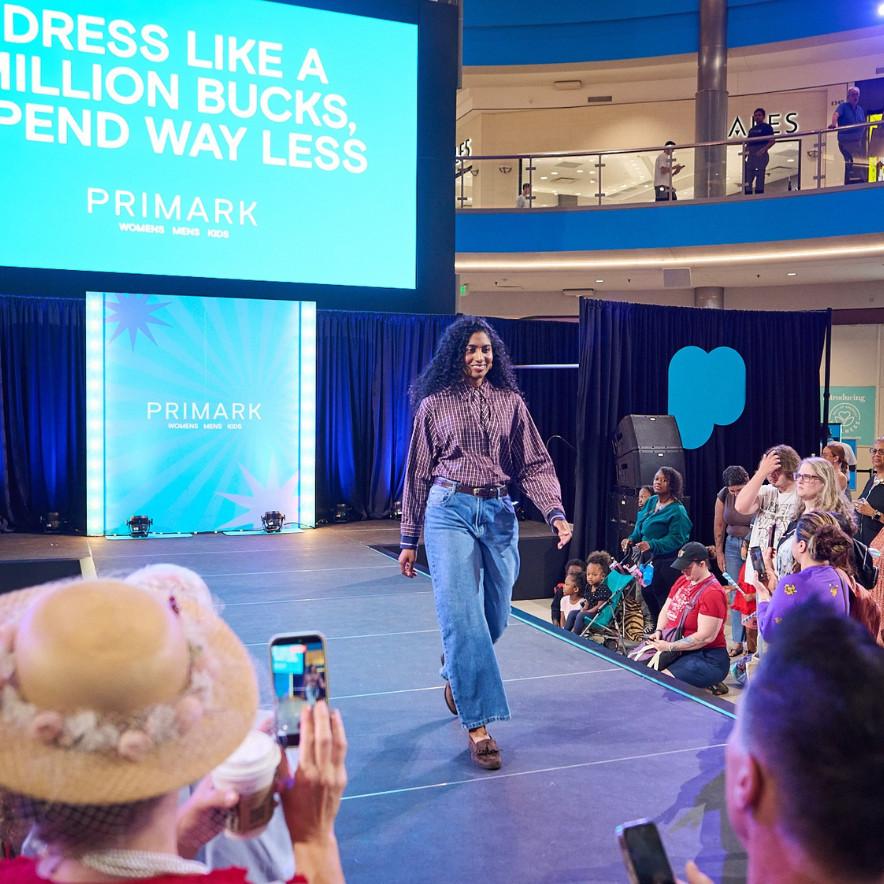 Model walks down a catwalk, surrounded by people taking pictures. Primark branding in background.