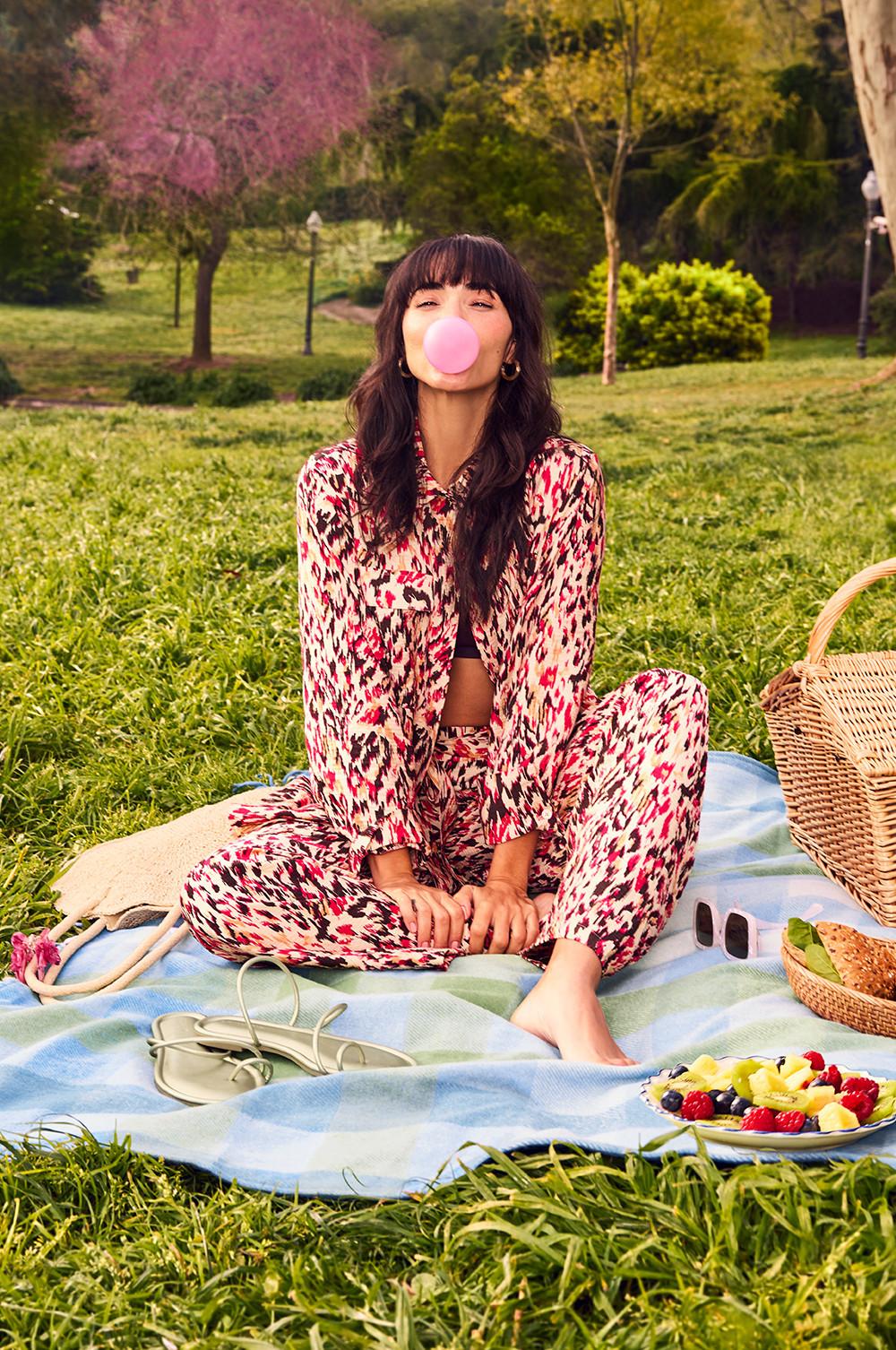 Person in einem rosa-schwarz gemusterten Kombiteil mit Palazzo-Hose, die auf einer blauen Decke mit einem Obstteller und rosa Kaugummi ein Picknick genießt.