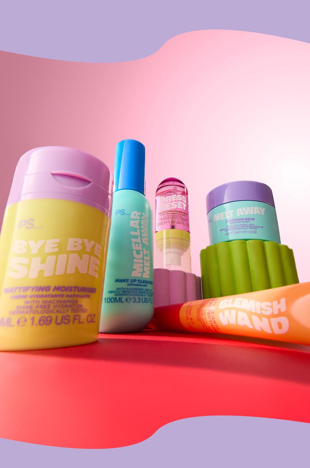 Assortimento di prodotti per la cura della pelle PS, tra cui la crema idratante Bye Bye Shine, acqua micellare detergente e l'applicatore per le imperfezioni su uno sfondo rosa.