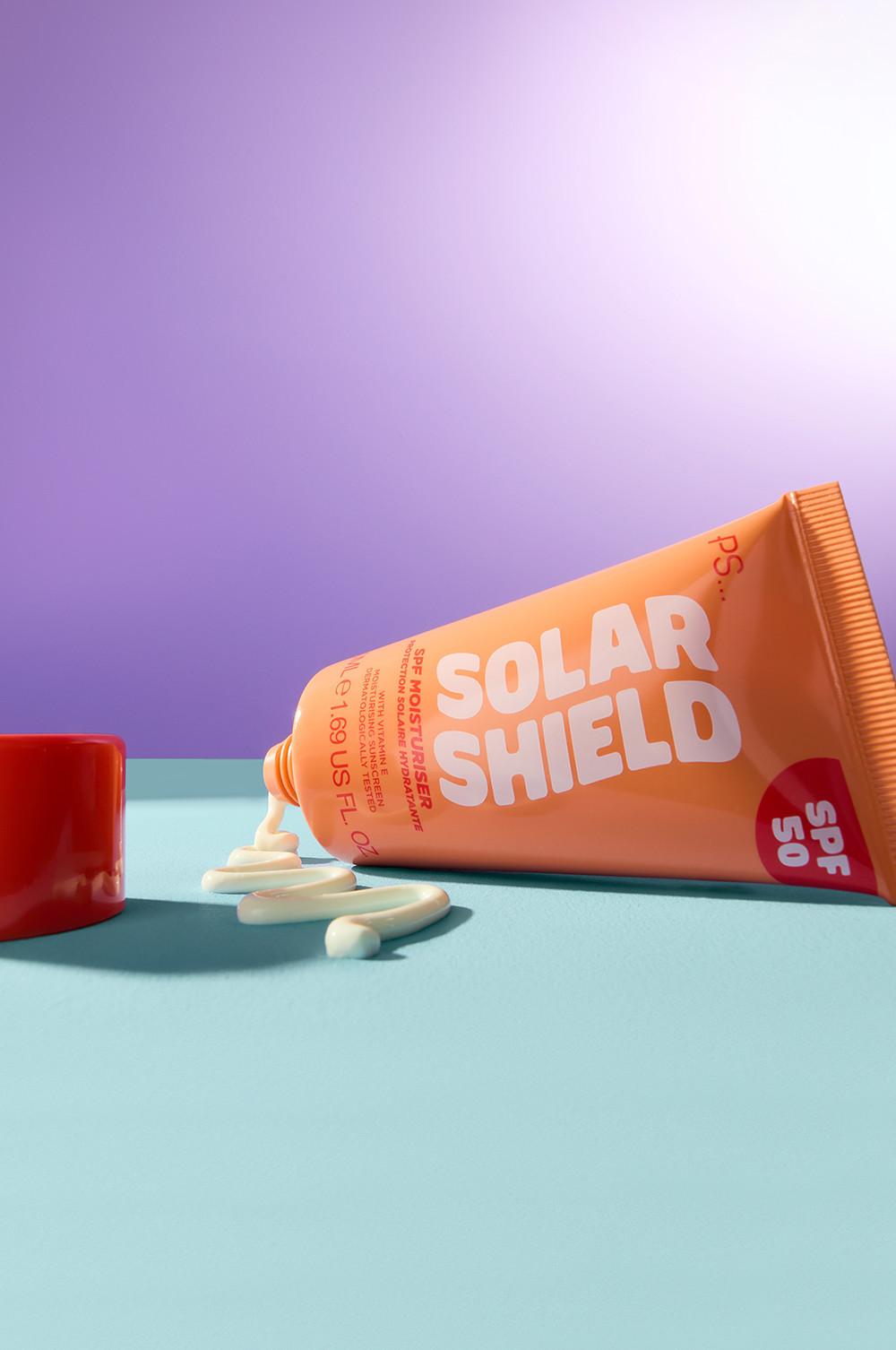 Tubetto di protezione solare Solar Shield arancione con SPF 50 e crema bianca spremuta fuori su una superficie turchese.