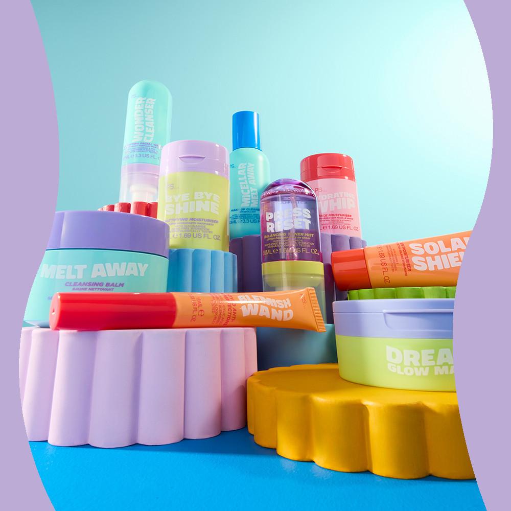 Assortimento di prodotti colorati per la cura della pelle, tra cui balsamo detergente, creme idratanti e sieri, disposti su piattaforme rosa e gialle su uno sfondo turchese.