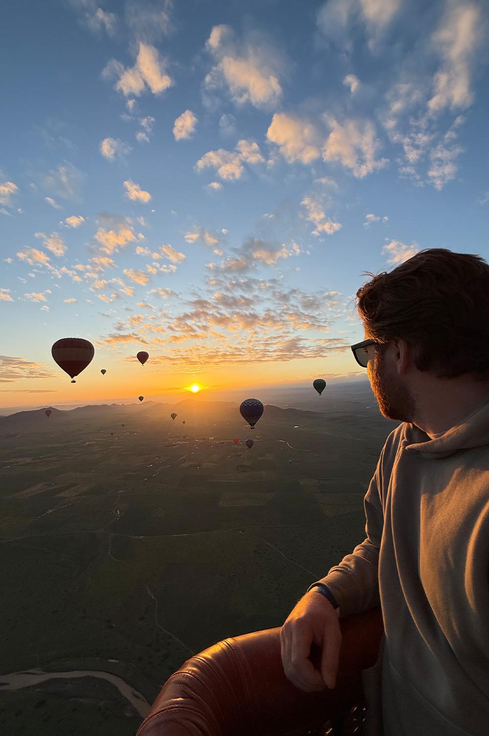 Personne dans une nacelle observant des montgolfières flotter au‑dessus du paysage au lever du soleil.