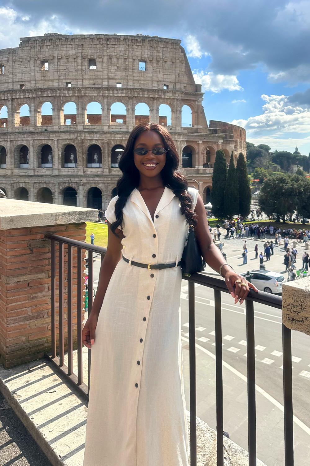 Mannequin portant une robe longue blanche ceinturée et des lunettes de soleil, posant à un belvédère avec le Colisée de Rome en arrière‑plan.