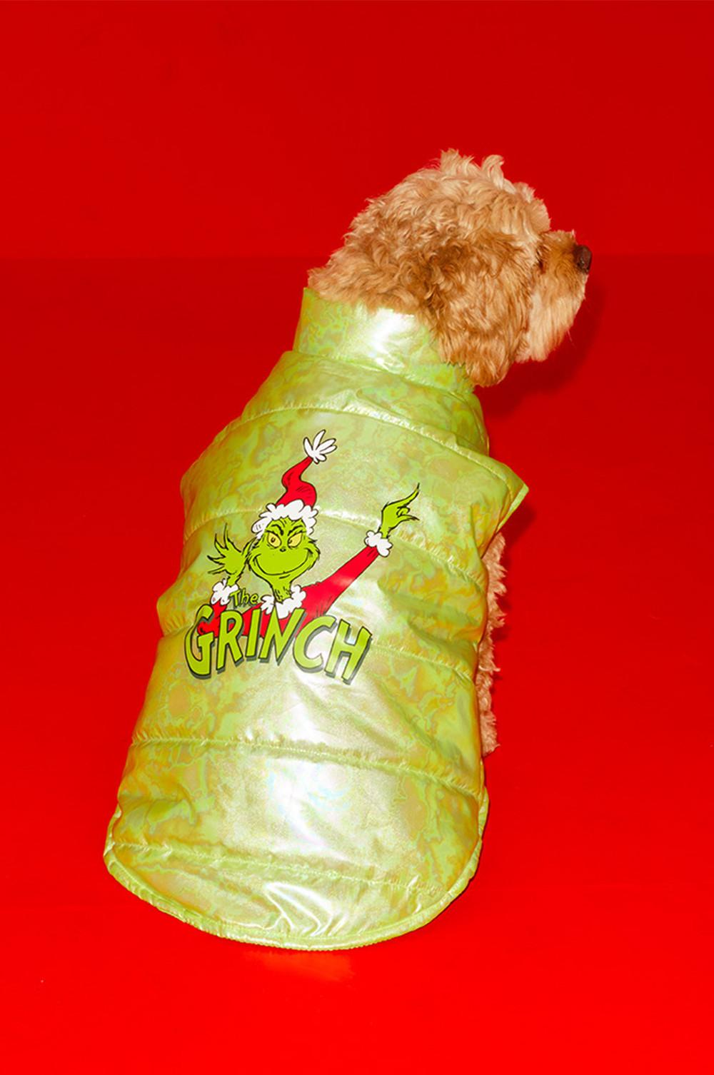 Small brown dog wearing a bright green padded coat featuring The Grinch character against a red background.