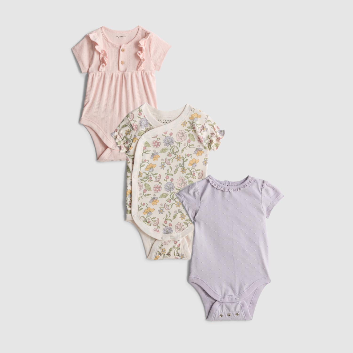 Three baby bodysuits: pink with ruffle trim, cream with floral print, and lilac with white spots.