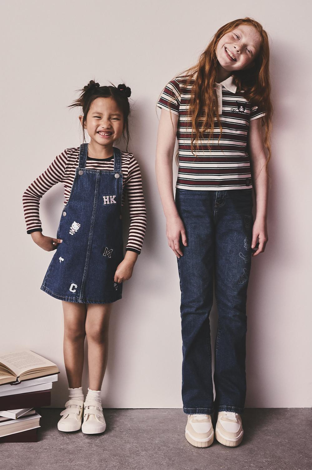 Due bambine sorridenti contro una parete neutra: una indossa maglietta a righe e jeans, l'altra un vestito a salopette con applicazioni.