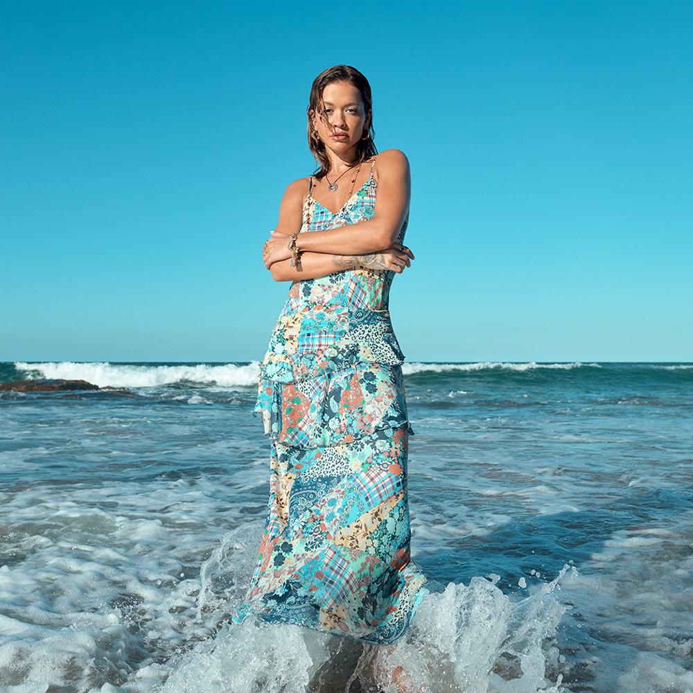 Rita Ora in piedi tra le onde dell'oceano mentre indossa un maxiabito patchwork con motivi floreali e scozzesi nei toni del blu e del corallo, sullo sfondo di un cielo azzurro terso.