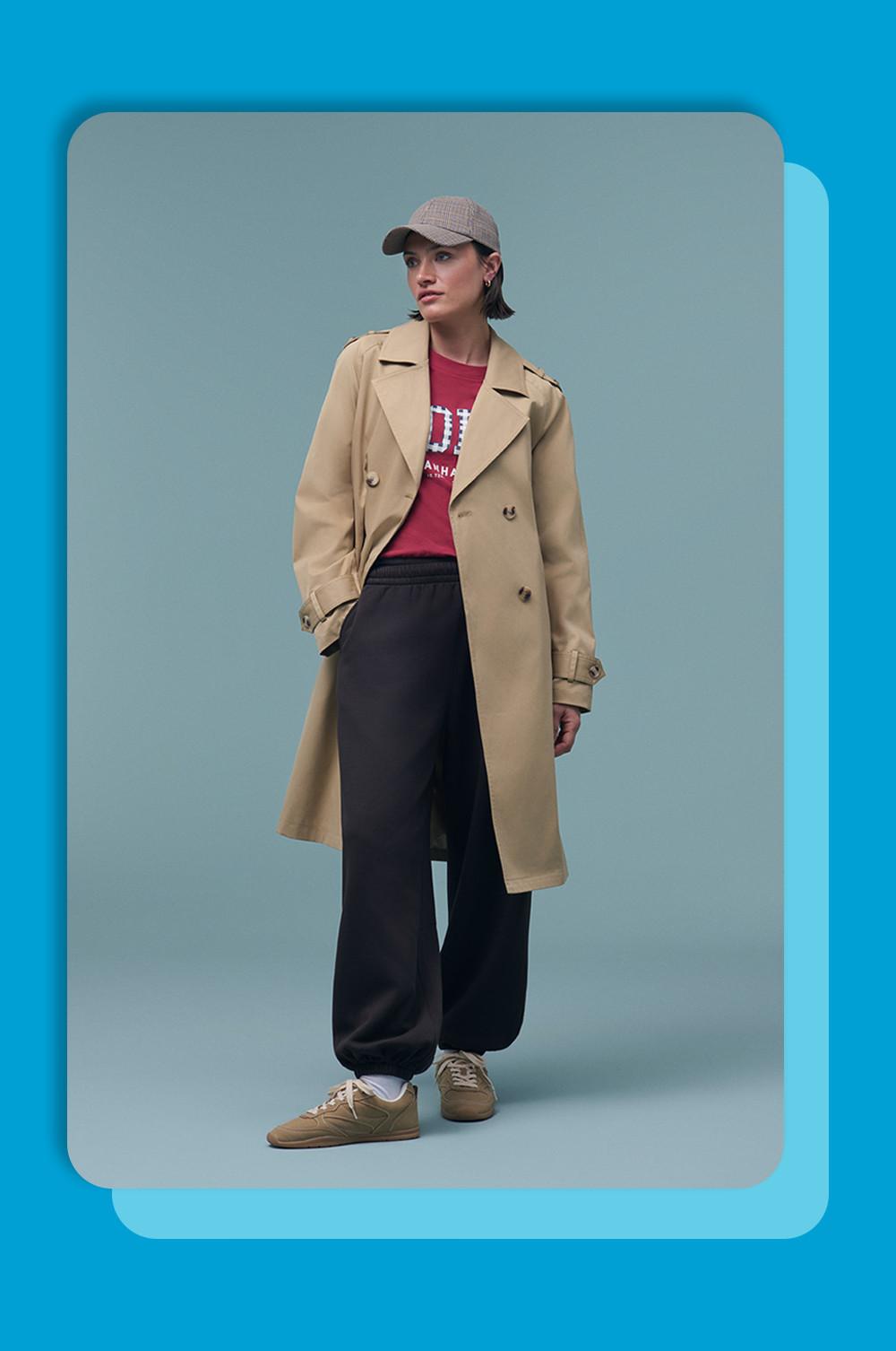 Model draagt een beige trenchcoat op een rood T-shirt met print en een zwarte broek met wijde pijpen, met een baseballpet en sneakers.