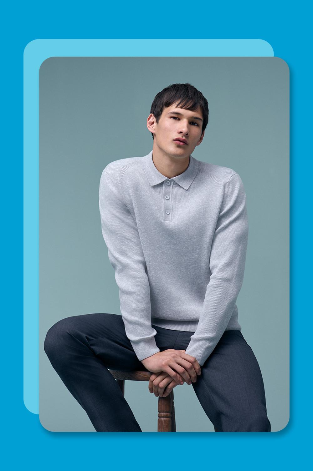 Model zit op een kruk en draagt een lichtgrijs gebreid poloshirt met lange mouwen op een donkere broek.