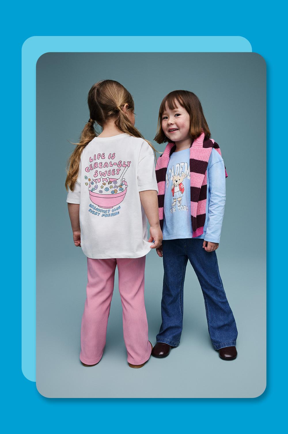 Twee meisjes in een T-shirt met print. De een draagt een roze broek en de ander een spijkerbroek en een gestreepte sjaal.