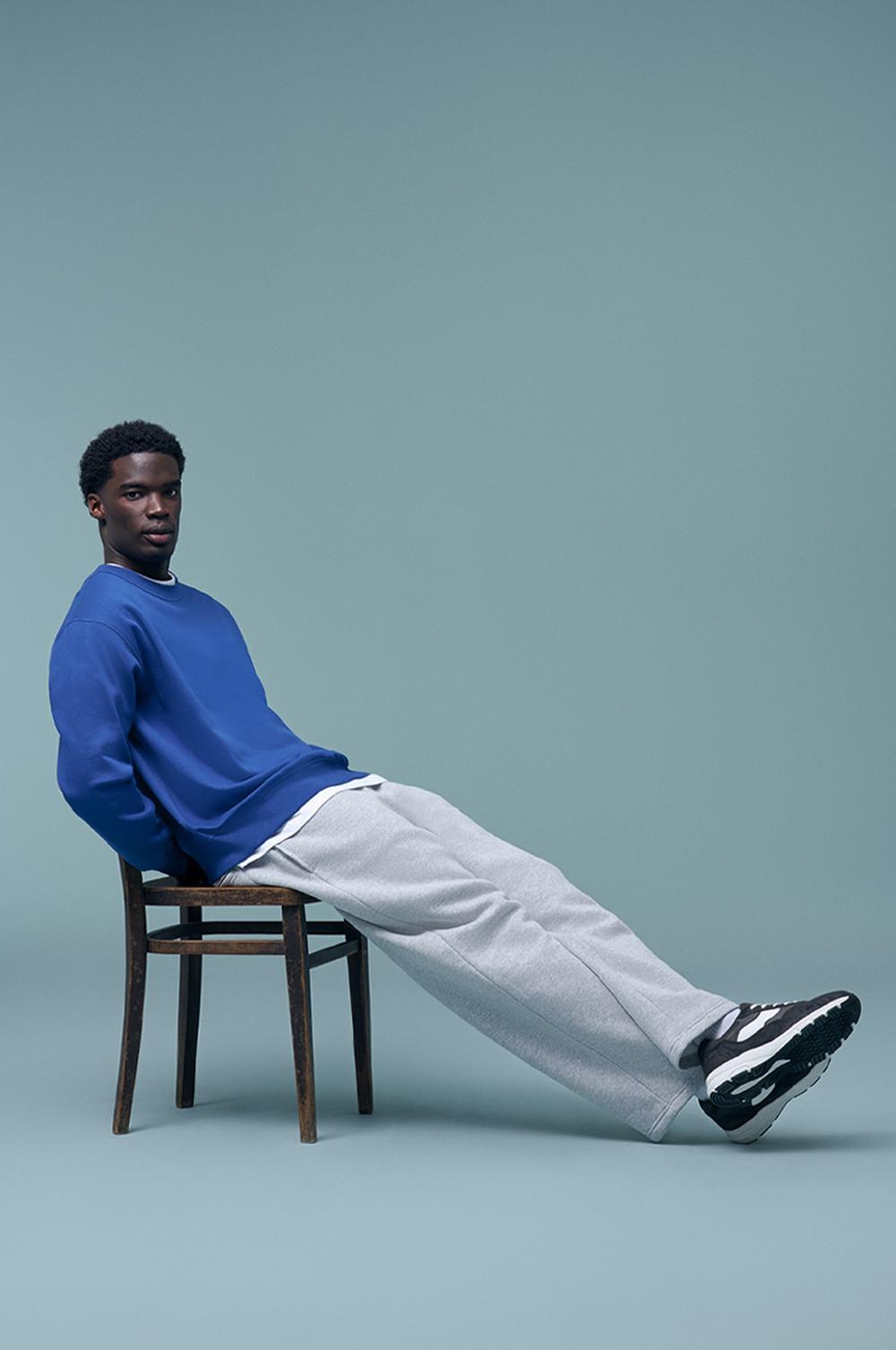 Model zit op een stoel en draagt een blauw sweatshirt, een grijze joggingbroek en zwart-witte sneakers.