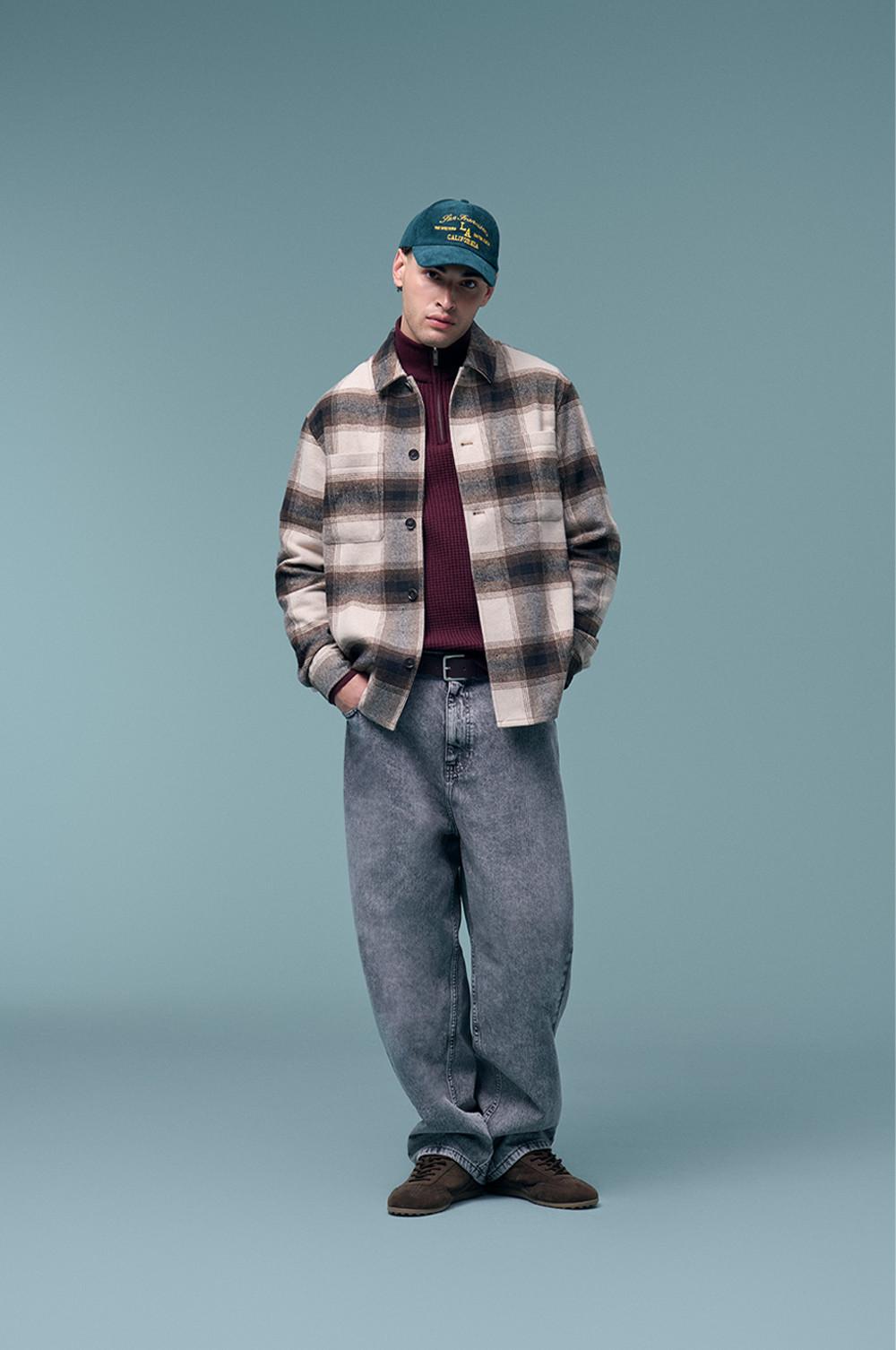 Model draagt een bruin- met roomwit geruit shacket op een bordeauxrode trui en een grijze spijkerbroek, en heeft een blauwgroene pet op.