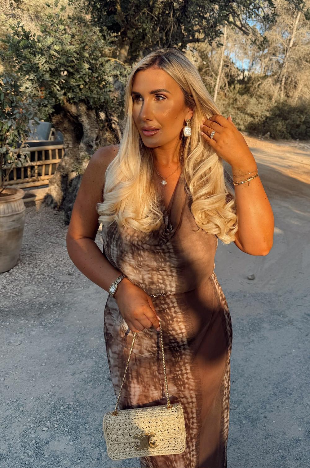 Person wearing a brown patterned dress with a woven straw handbag outdoors in a garden setting.