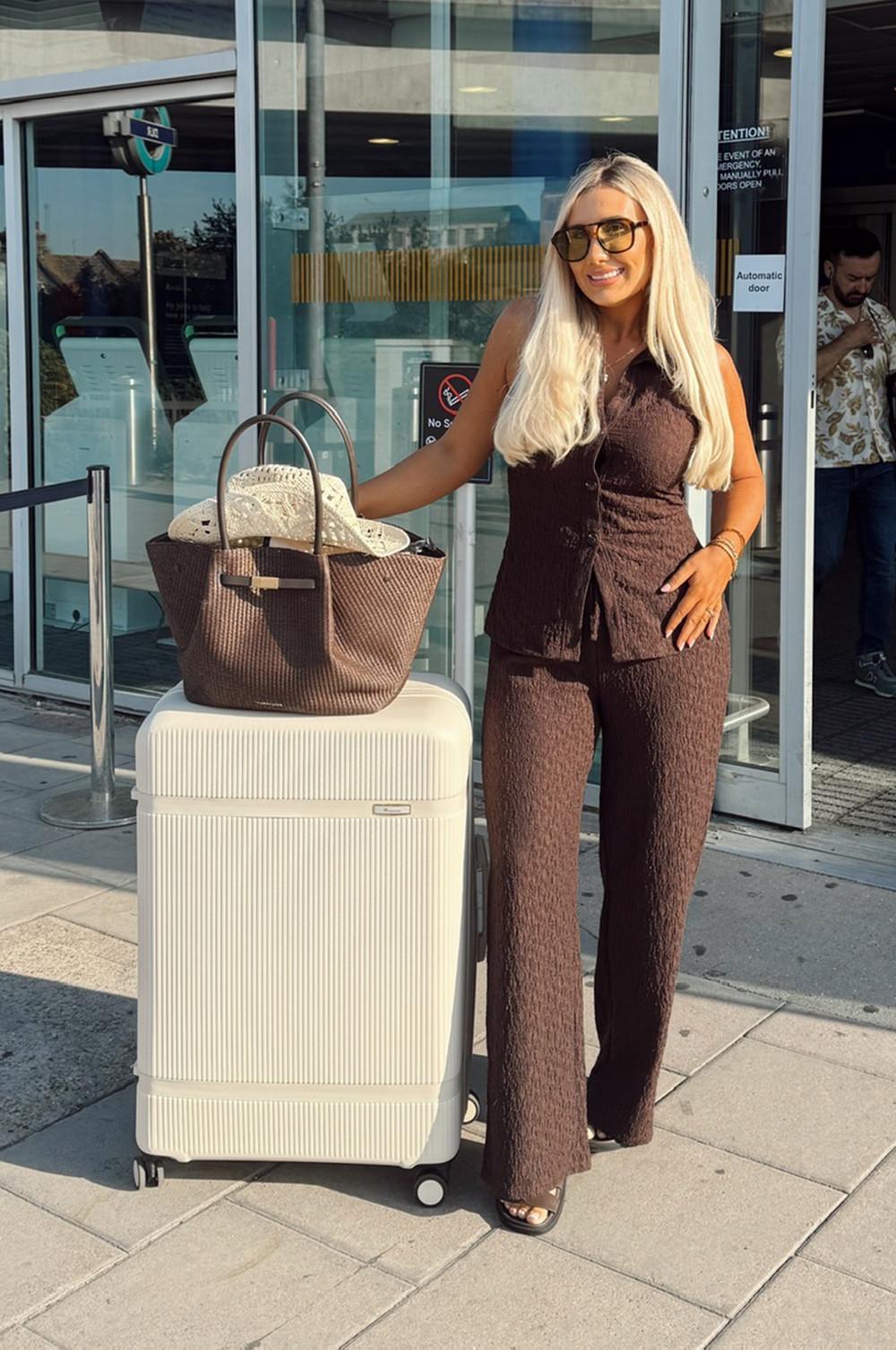 Person in brown outfit standing beside cream suitcase with straw hat in woven brown tote bag outside a building entrance.
