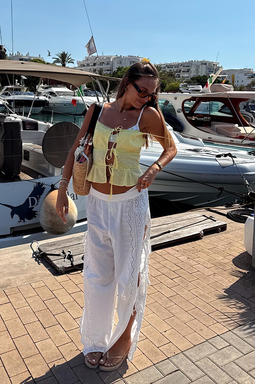 Person in yellow crop top and white eyelet pants standing on a marina dock with boats in the background.