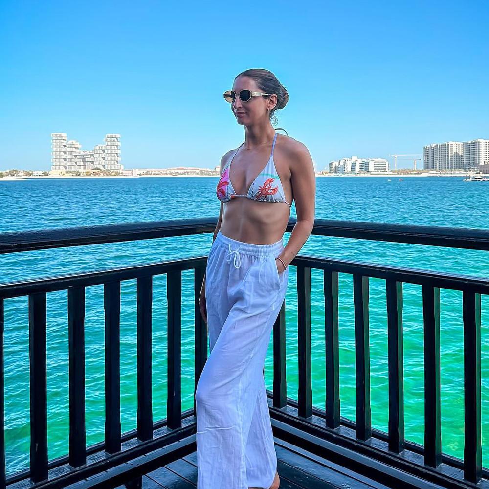 Person in printed bikini top and white beach trousers enjoying coastal view from balcony.