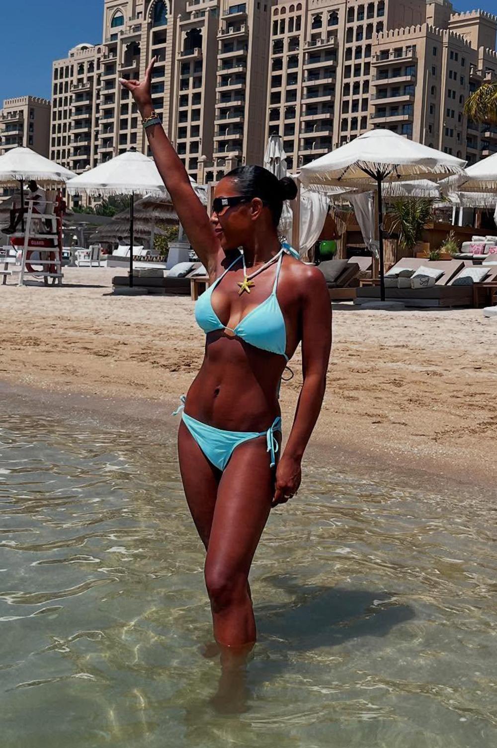 Person in light blue bikini standing in shallow water at a beach resort with white umbrellas and luxury buildings in background.