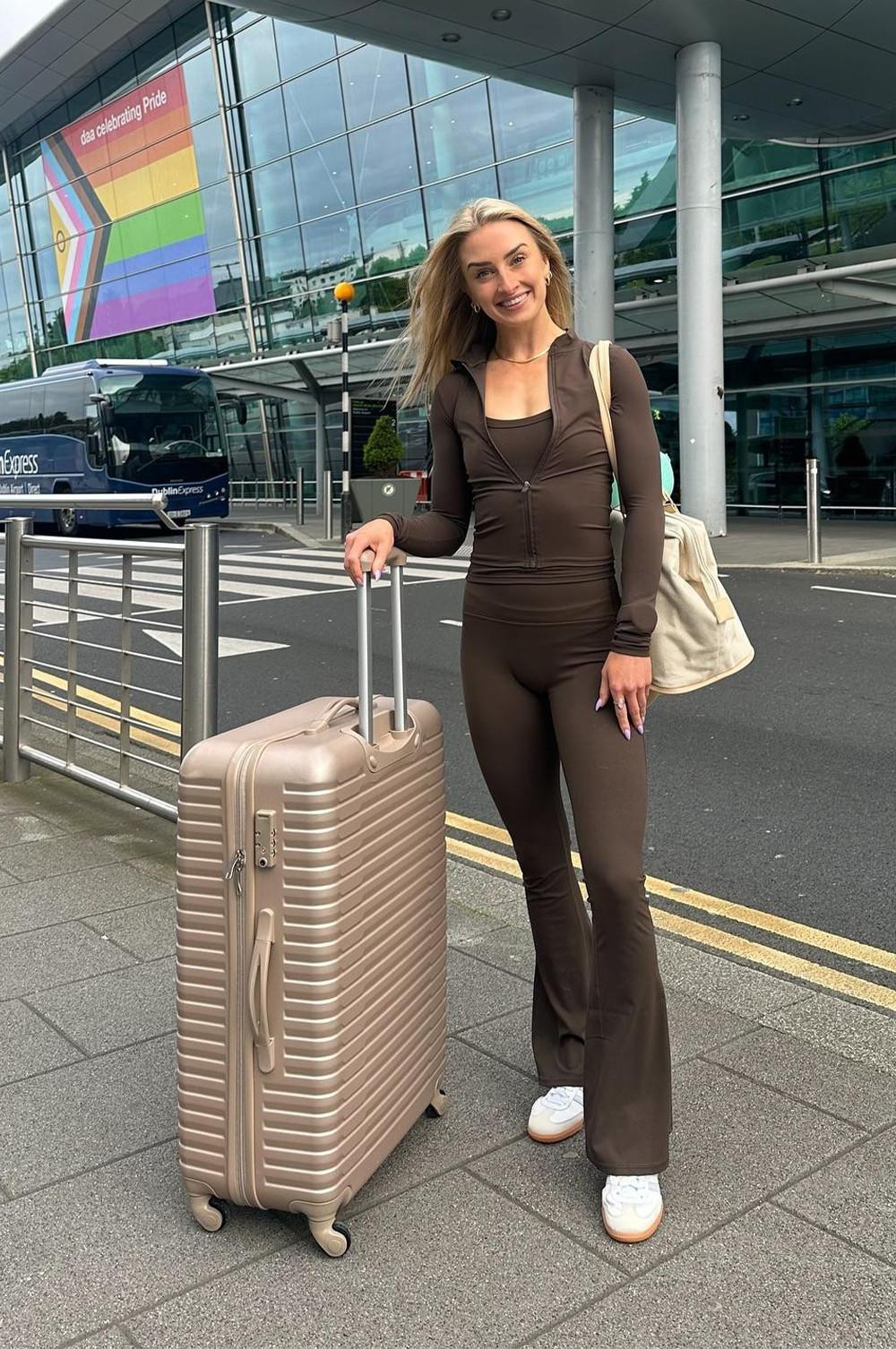 Brown fitted jumpsuit with cream shoulder bag and rose gold suitcase at airport. Pride flag visible in background.