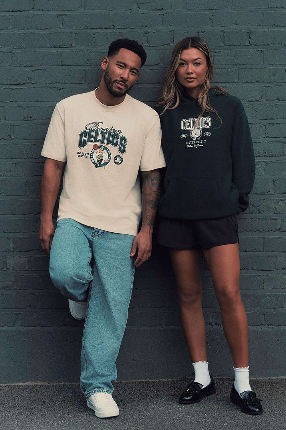 Dos modelos delante de una pared de ladrillo verde azulado con una camiseta de color crema y una sudadera con capucha de los Boston Celtics, respectivamente.
