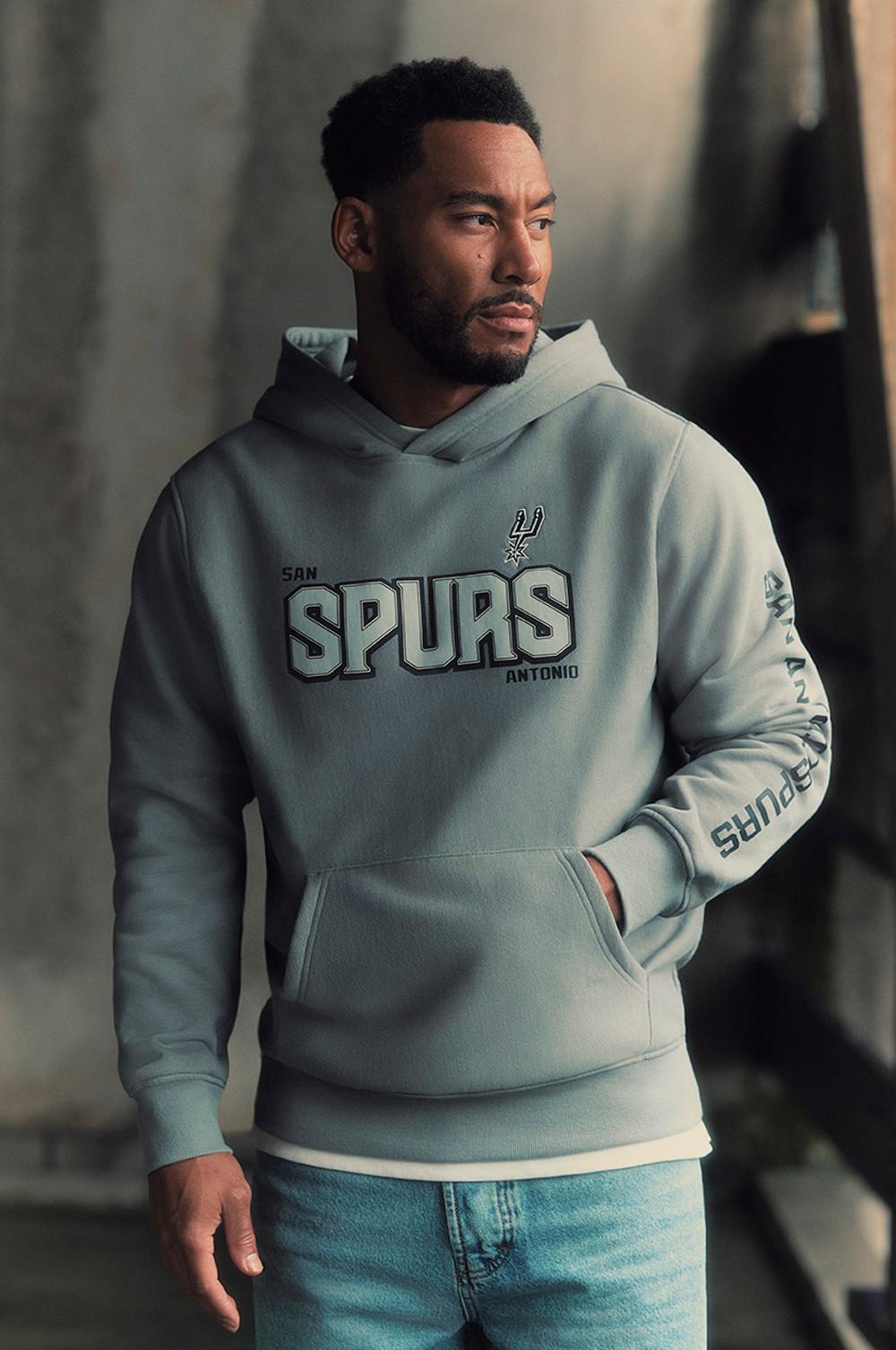 Sudadera con capucha gris claro de los San Antonio Spurs con el logotipo del equipo y texto en la manga, combinada con unos tejanos azules.