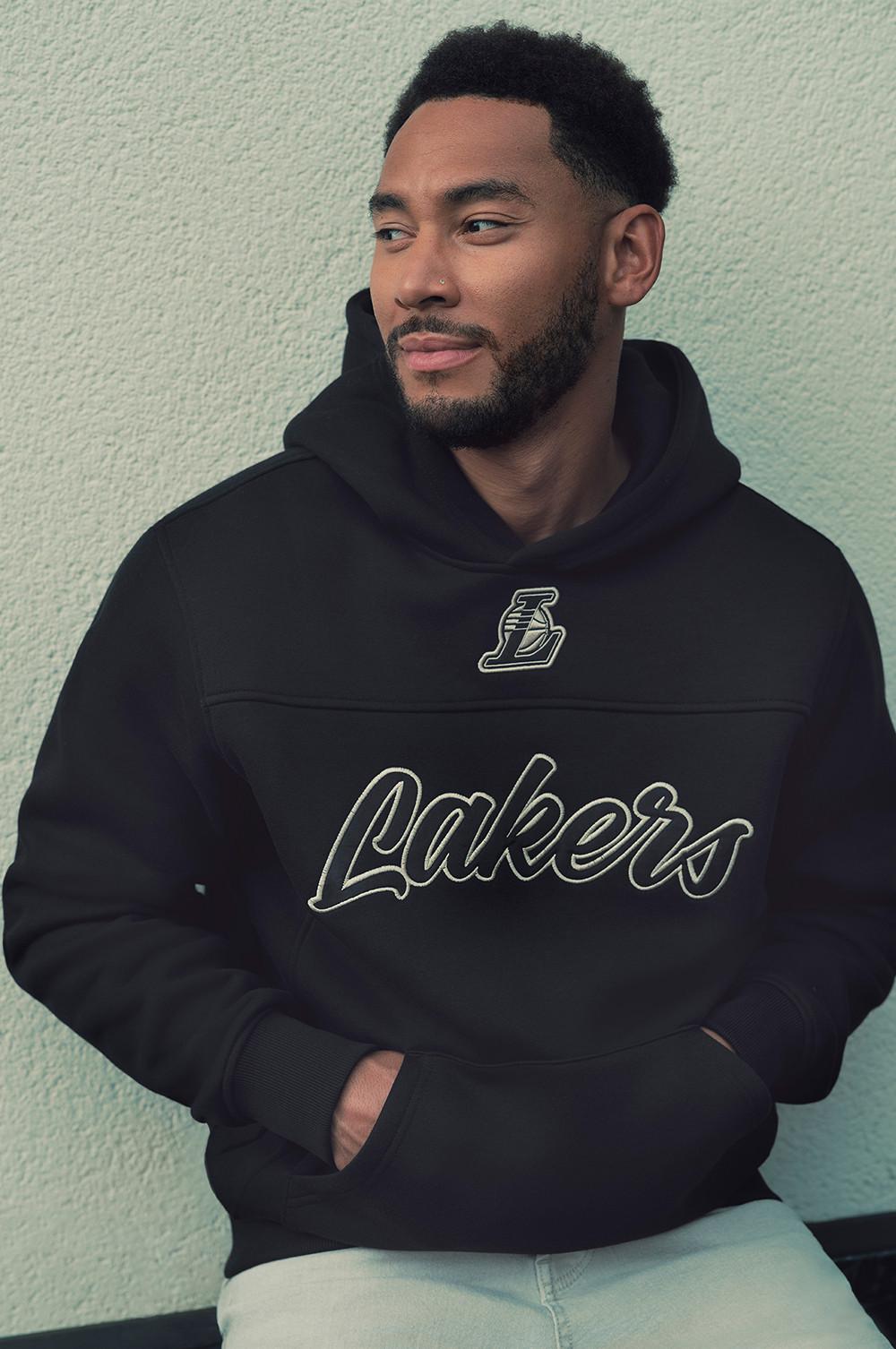 Modelo con sudadera con capucha negra de los Lakers con el logotipo del equipo y letras delante una pared de color claro.
