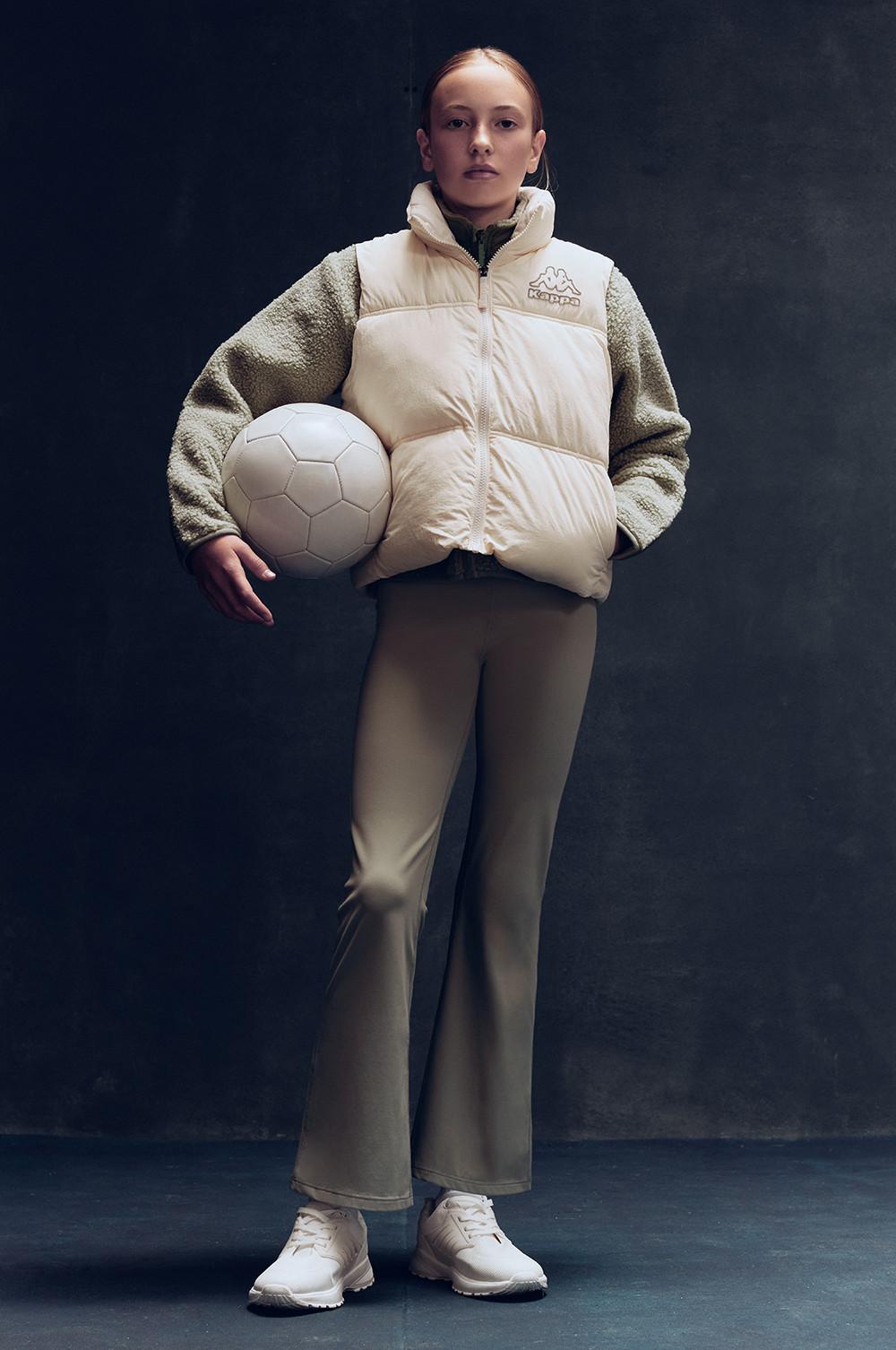 Persona con giacca e pantaloni panna che tiene in mano un pallone da pallavolo bianco e indossa scarpe da ginnastica bianche.