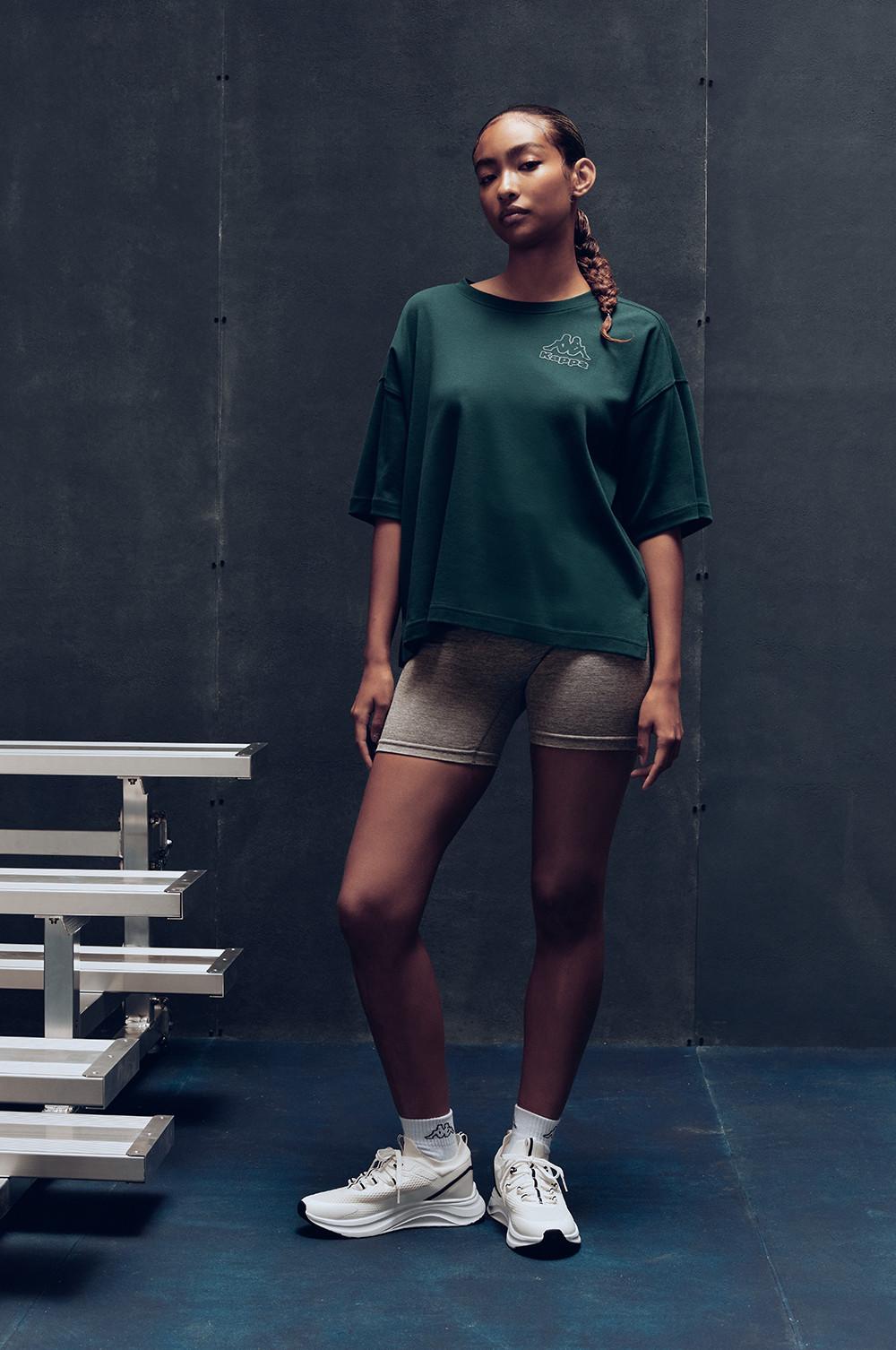 Persona che indossa una maglietta oversize verde con pantaloncini, collant bordeaux e scarpe da ginnastica bianche in piedi vicino a una scala di legno.