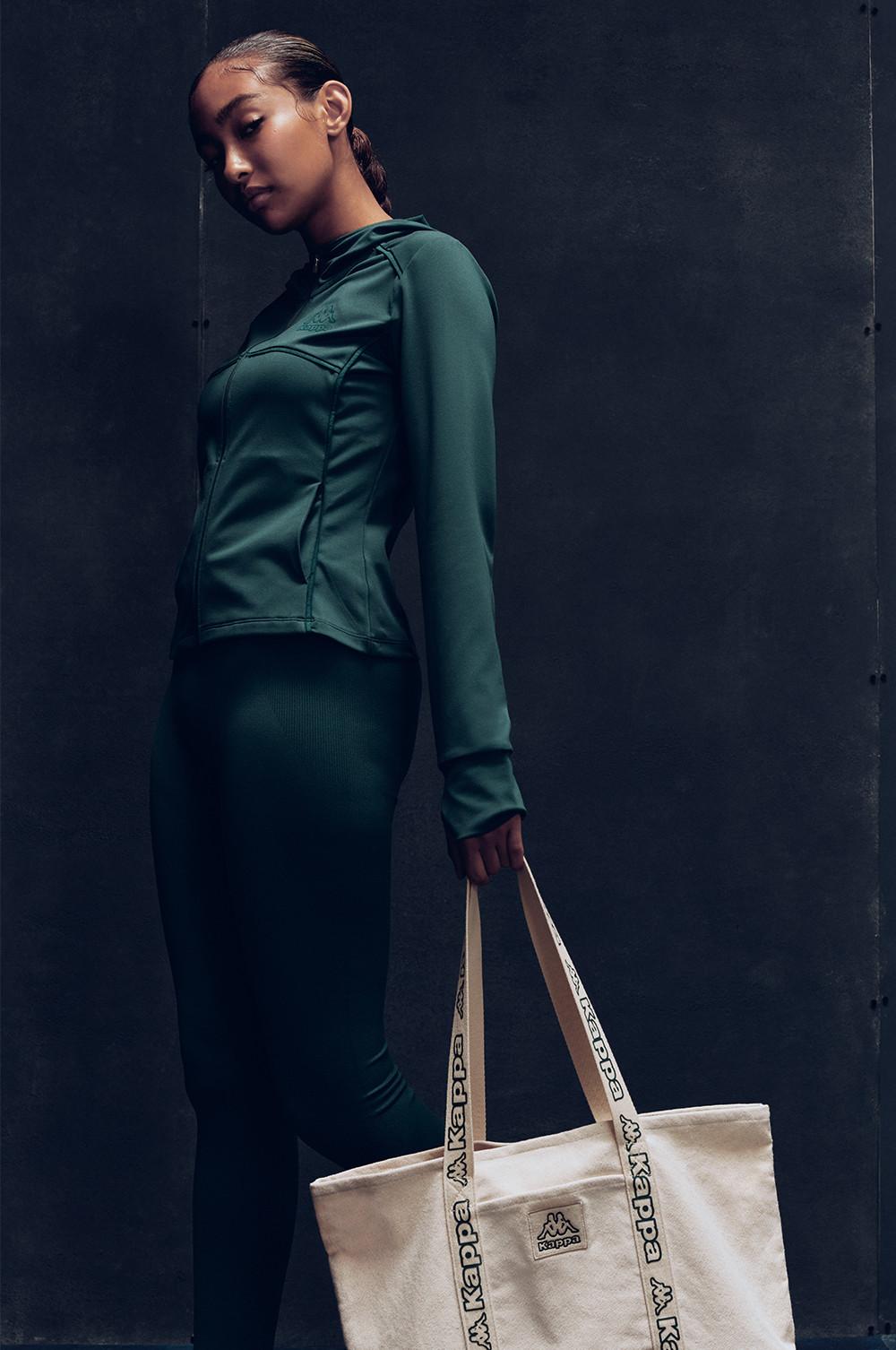 Persona che indossa un completo verde scuro e tiene in mano una borsa tote panna con manici in corda su sfondo scuro.