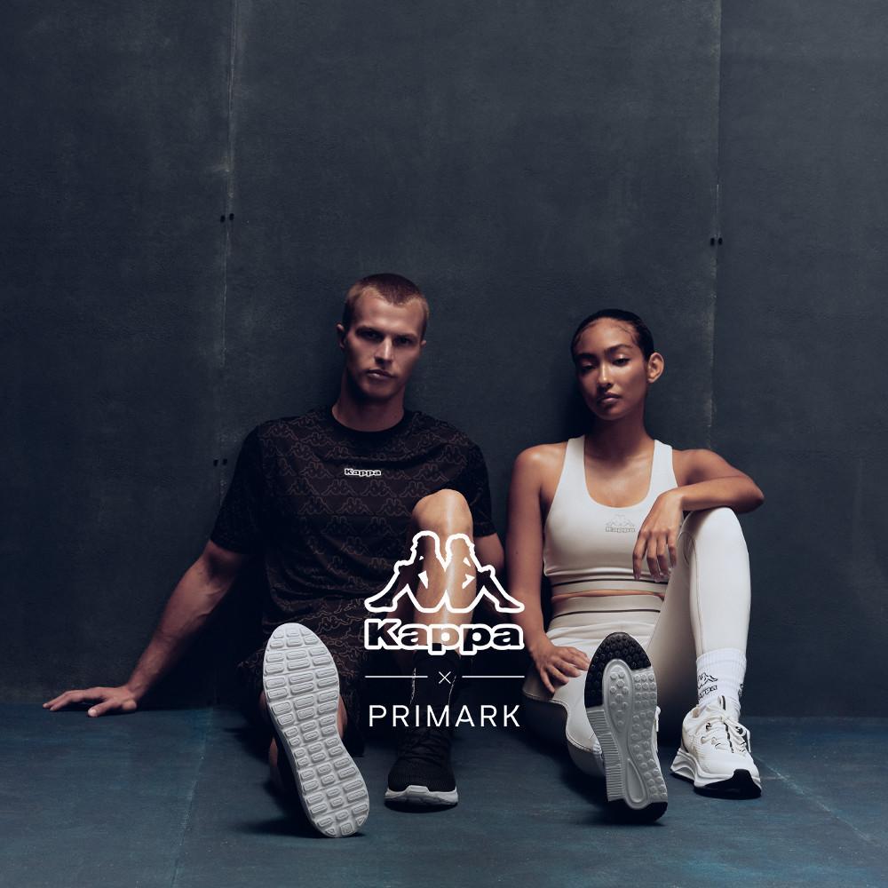 Due persone sedute contro una parete scura che indossano capi della collezione di abbigliamento sportivo Kappa x Primark.