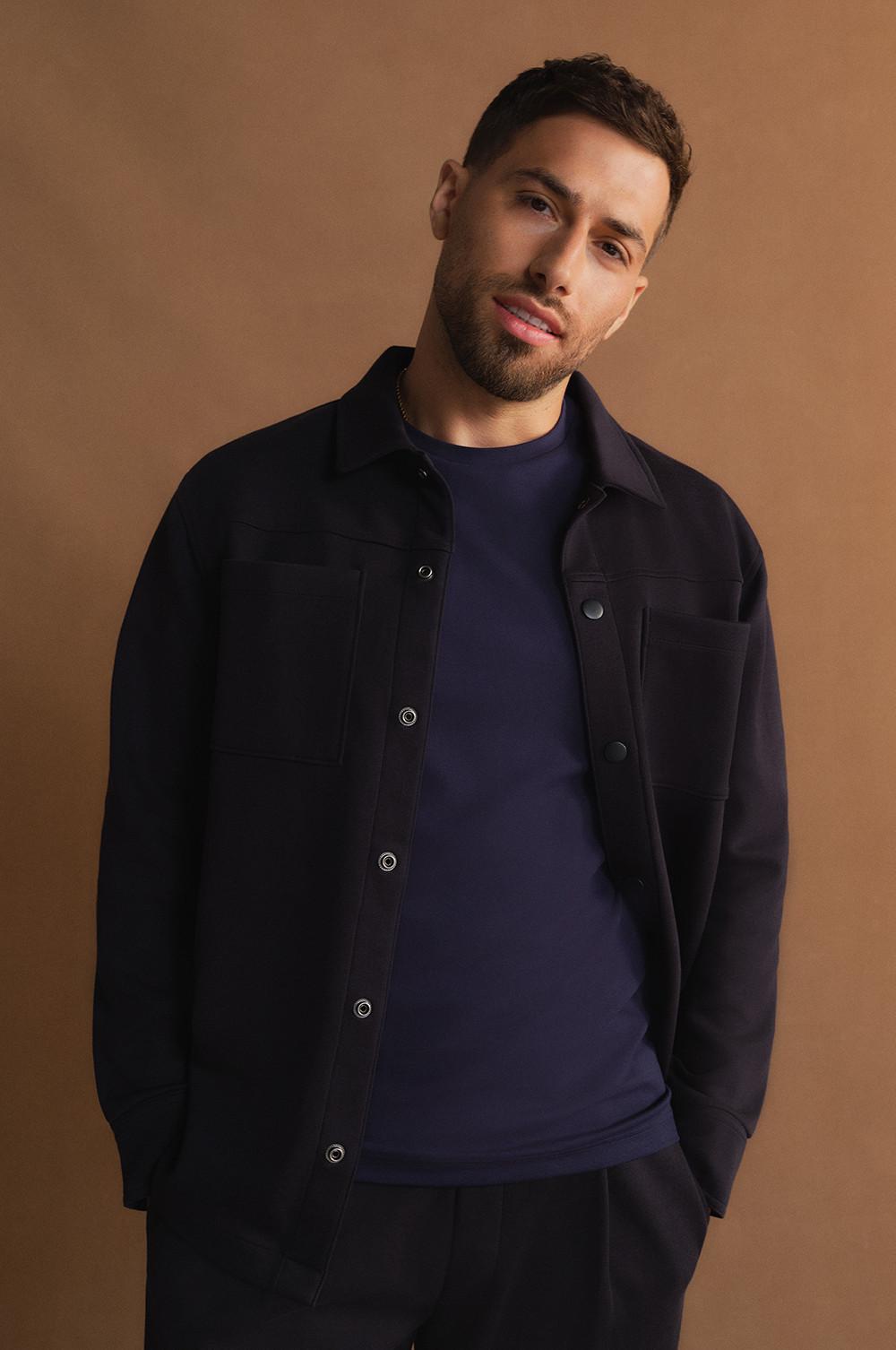 Personne portant une surchemise noire à poches, sur un t-shirt bleu marine, le tout sur fond beige chaud.