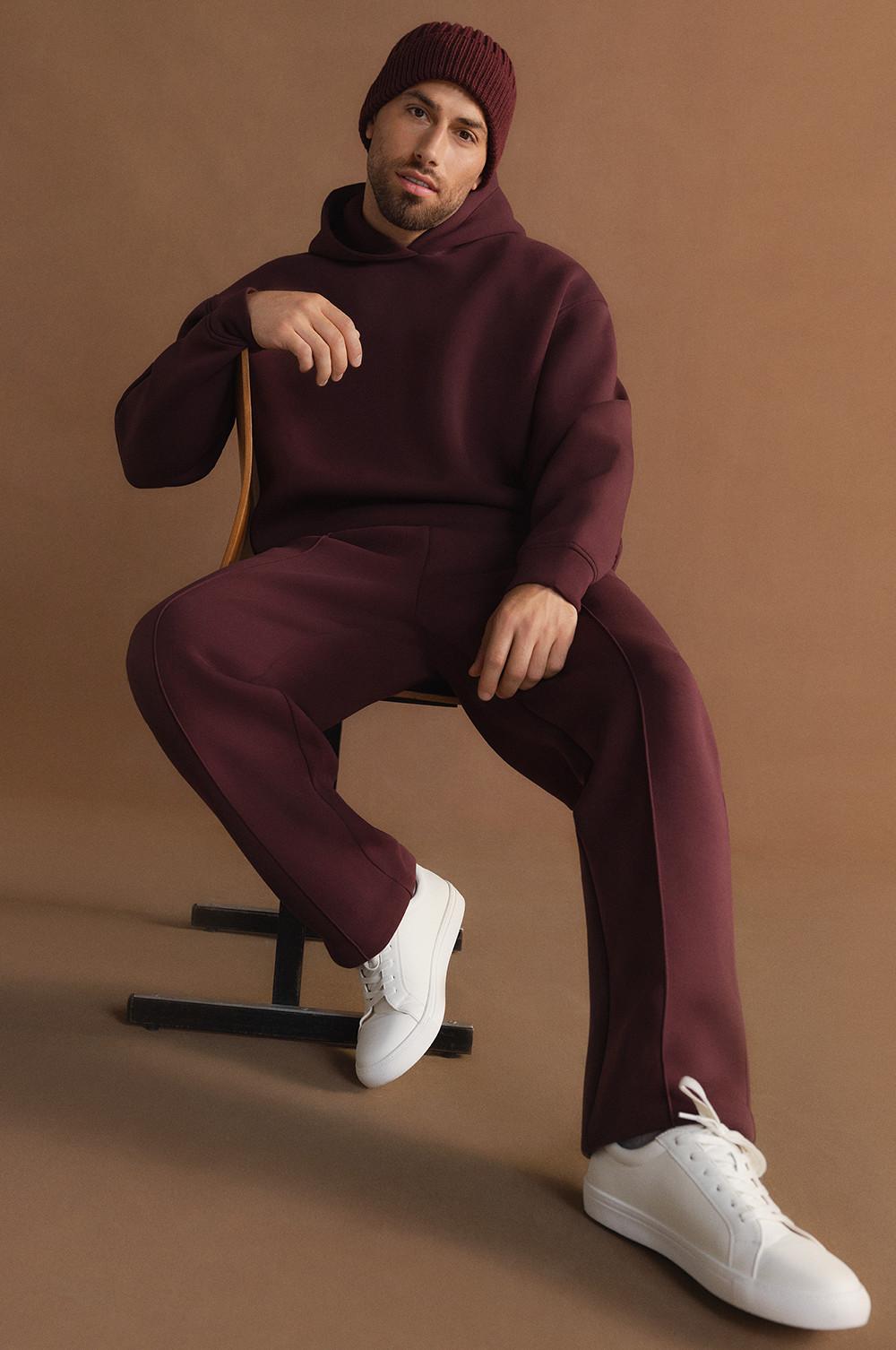 Personne portant un survêtement bordeaux avec bonnet assorti et baskets blanches, assise sur une chaise.