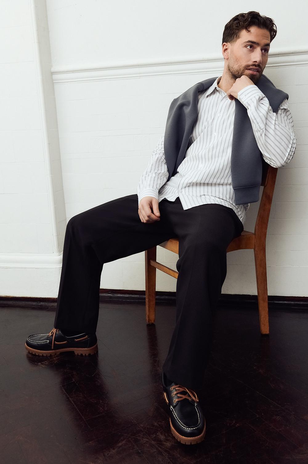 Kem wearing a striped shirt, grey jumper over the shoulders and black trousers with black shoes