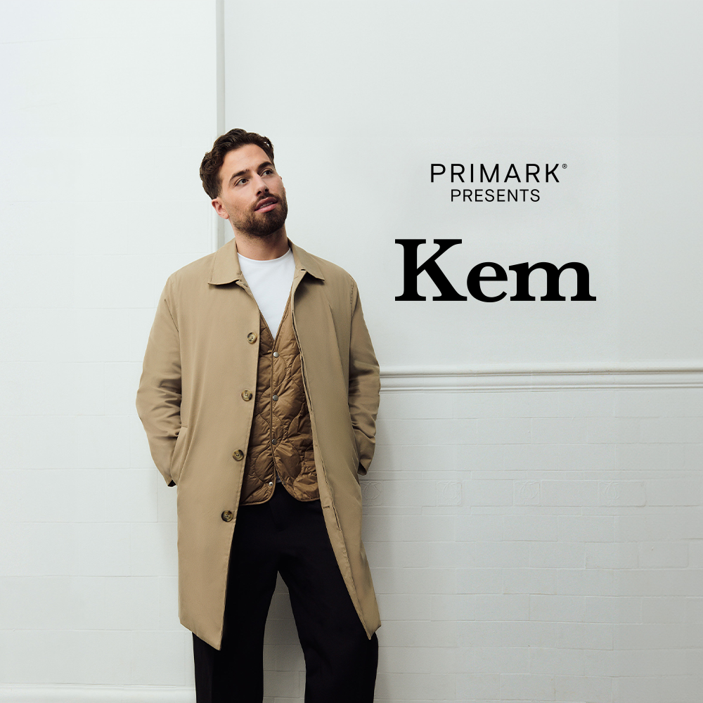 Kem Cetinay wearing a beige trench coat, black trousers, brown jacket under the trench coat and white t-shirt