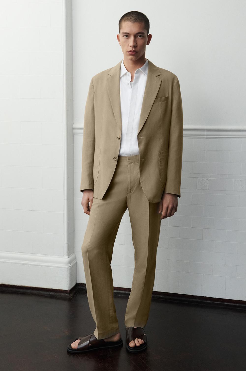 Man wearing beige suit, white shirt, and brown sandals standing indoors.