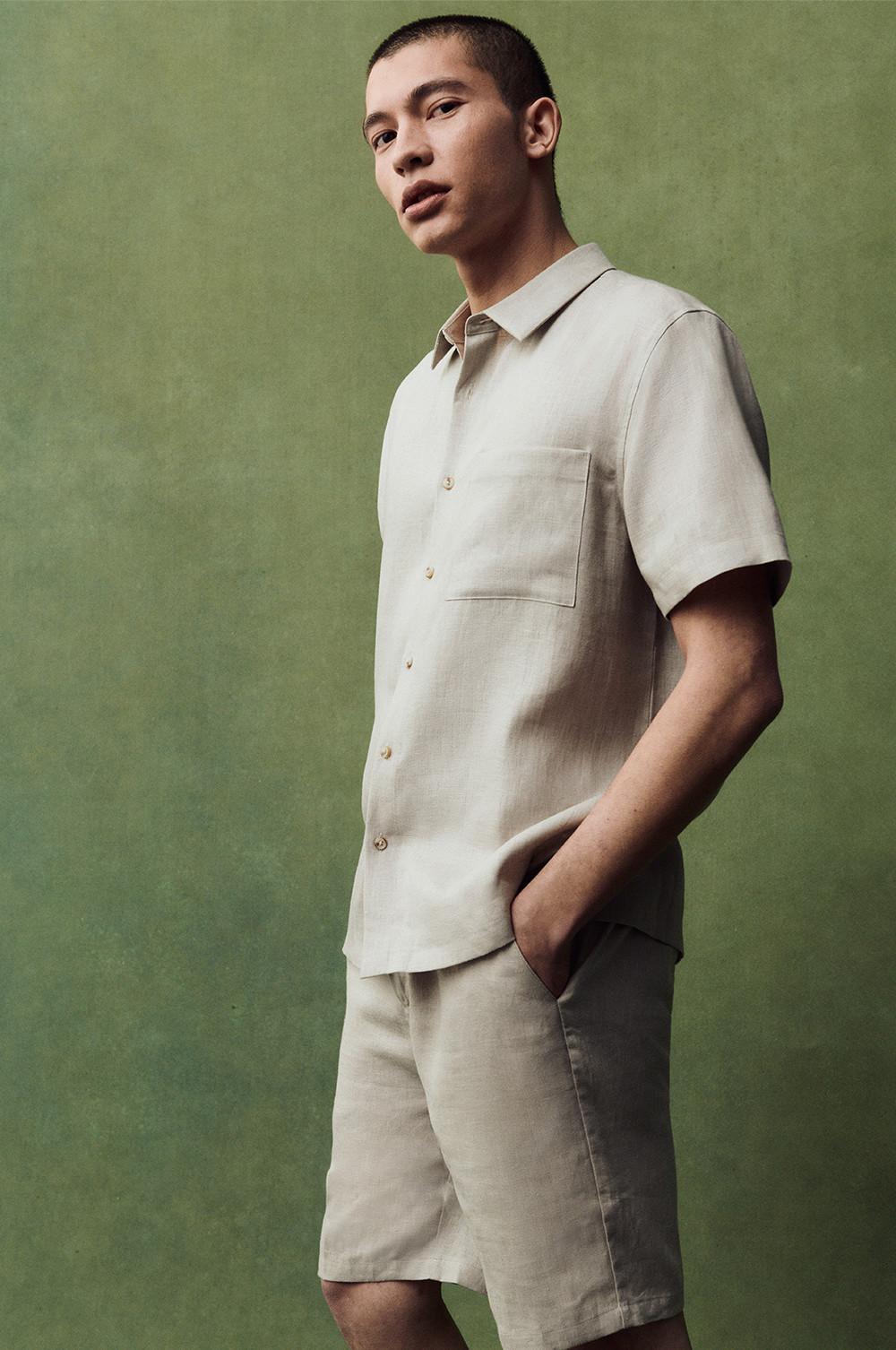 Model in beige short-sleeve shirt and matching shorts, hands in pockets, against a green background.
