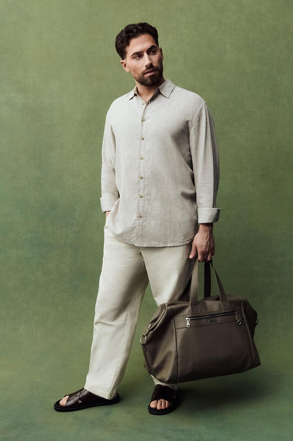 Kem wearing beige linen shirt with cream trousers, holding brown leather weekend bag against green background.