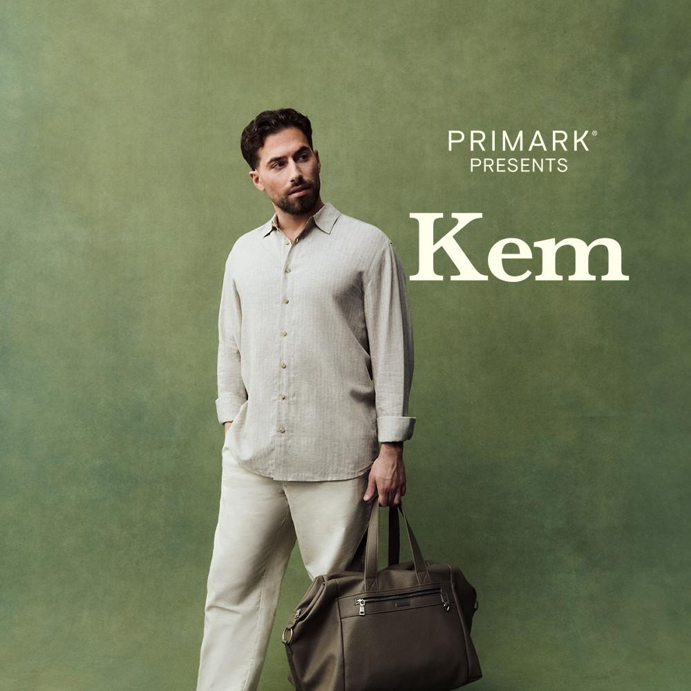 Kem Cetinay wearing beige linen shirt with light trousers and leather sandals, holding a brown leather travel bag against a green background.