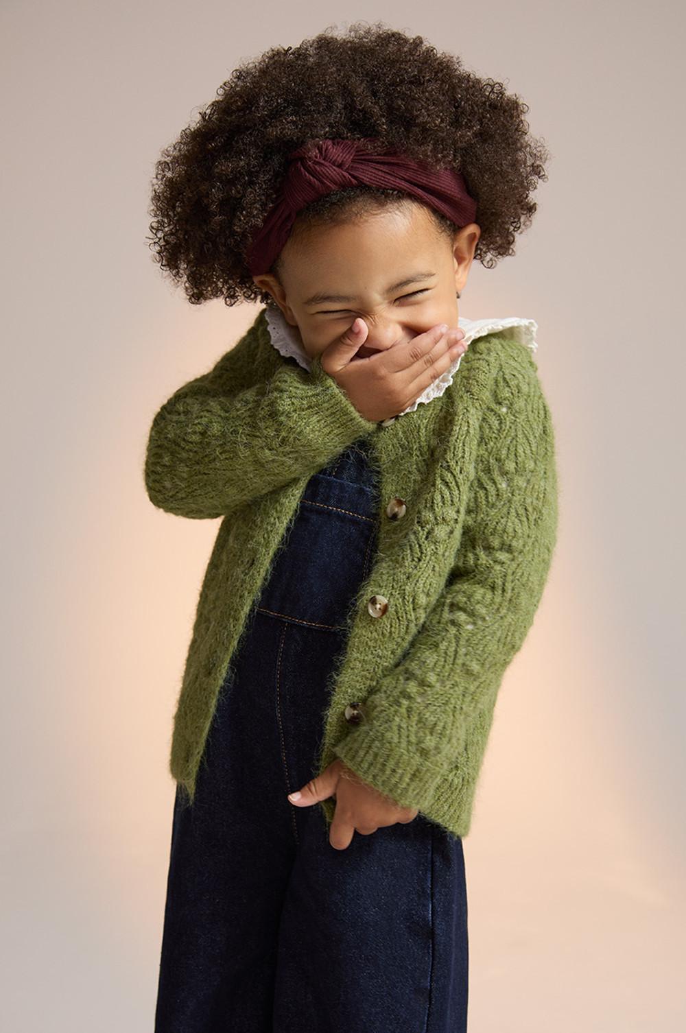 Child wearing green cable knit cardigan over denim dungarees with burgundy headband, covering mouth while laughing.