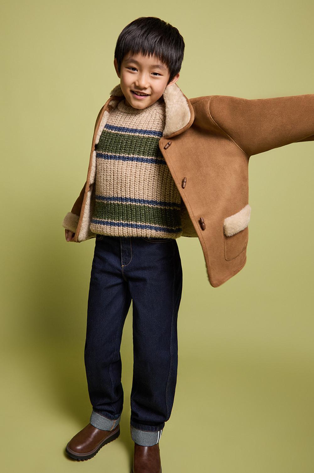 Child wearing striped knitted jumper, dark jeans and brown coat with fleece lining against light green backdrop.