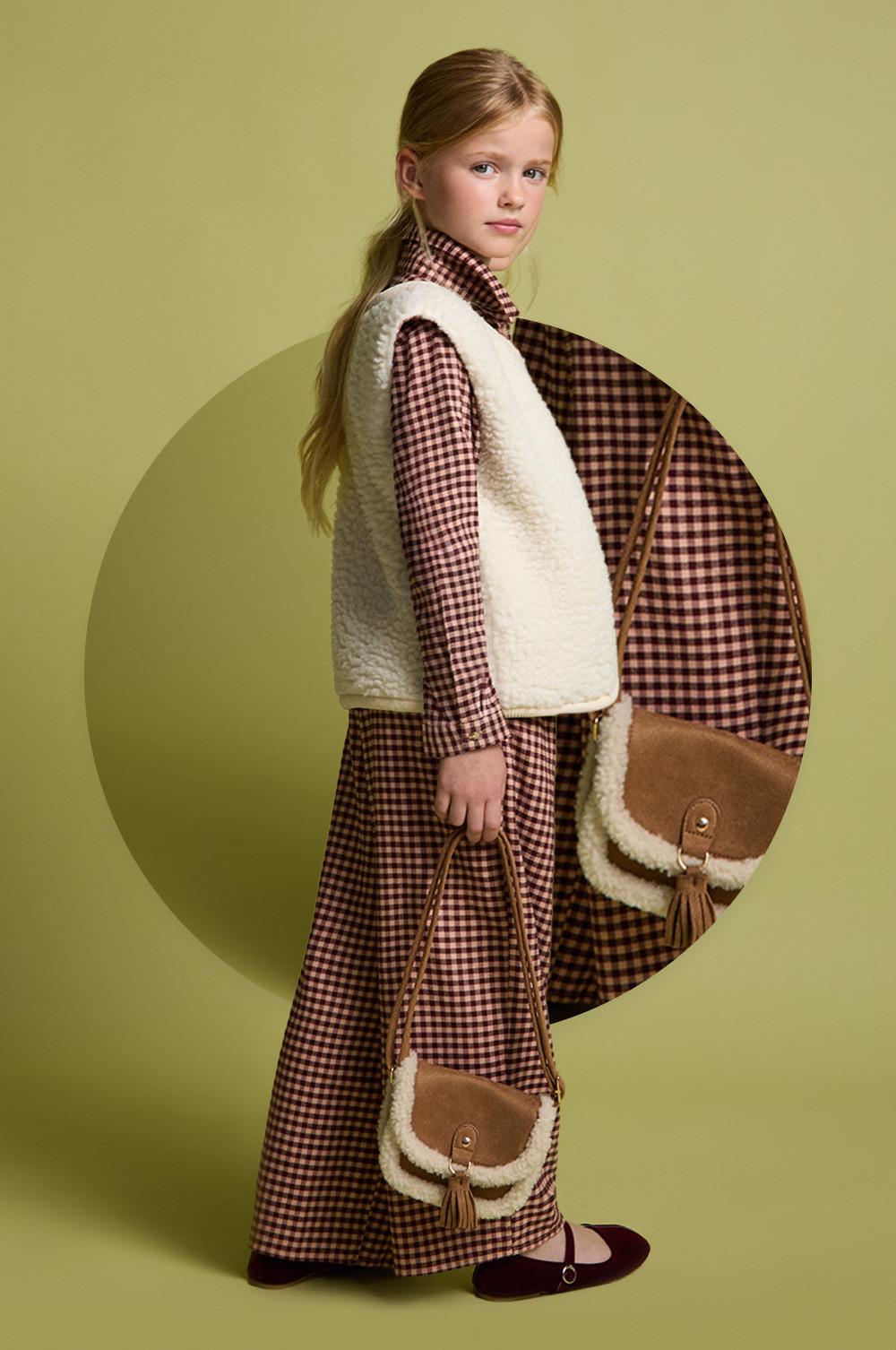 Child wearing checkered dress with cream sherpa gilet and matching brown crossbody bag against green background.
