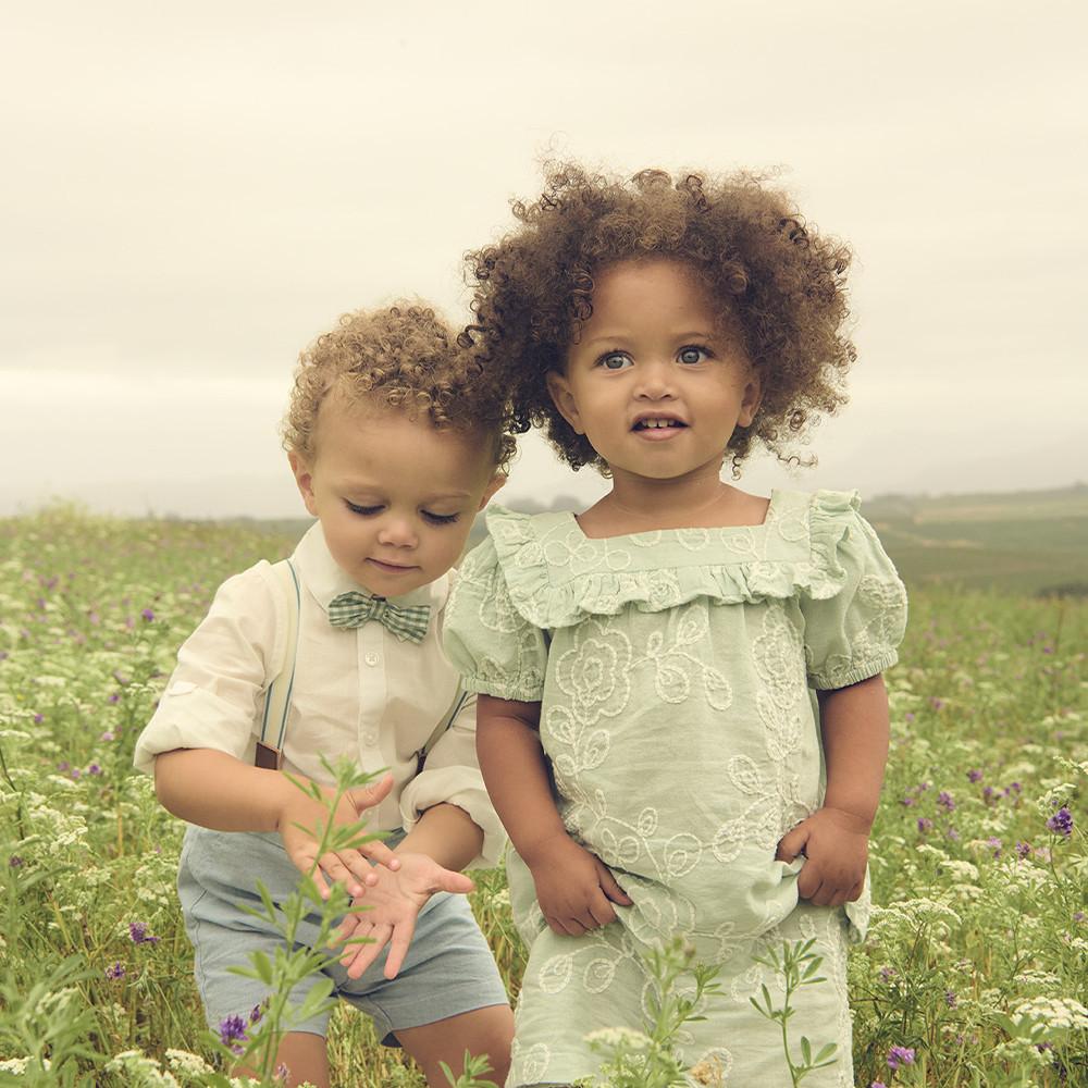 Due bambini in abiti eleganti su un prato fiorito. Il bimbo indossa papillon e pantaloncini, la bimba un vestito verde chiaro ricamato.