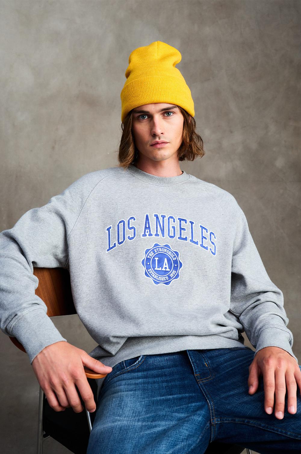 Model wears Los Angeles sweatshirt in grey, paired with yellow beanie and jeans