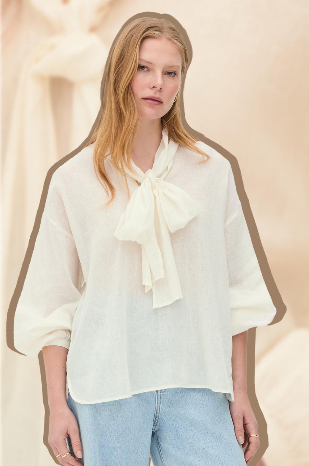 Model wearing a cream blouse with long sleeves and bow tie, paired with light blue jeans.