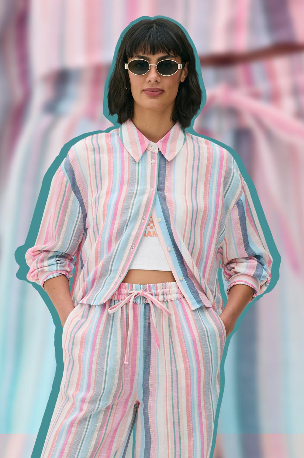 Woman in striped shirt and trousers with sunglasses and hands in pockets.