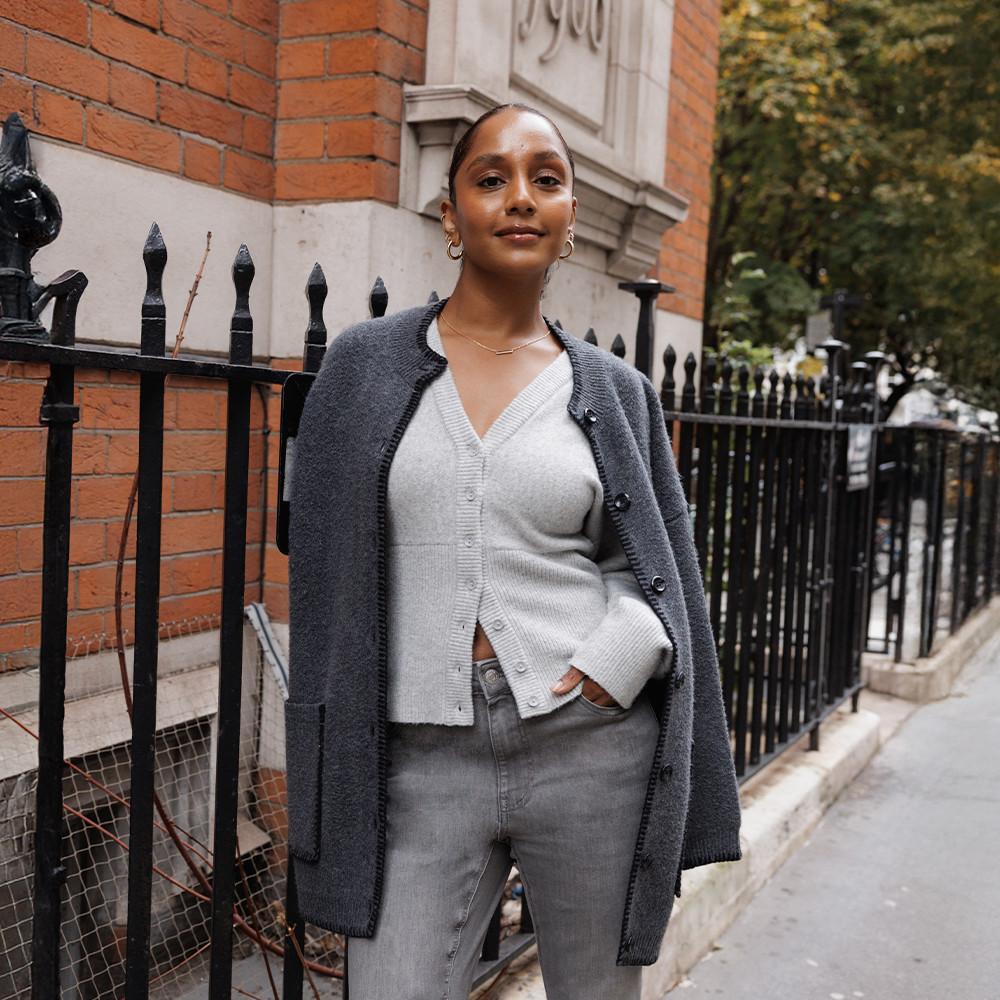 Grey cardigan layered with charcoal overcoat and light grey jeans, standing beside brick building.