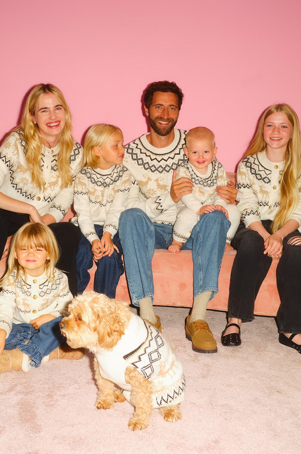 Family wearing matching Nordic patterned jumpers sitting together against pink background, dog in coordinating outfit.